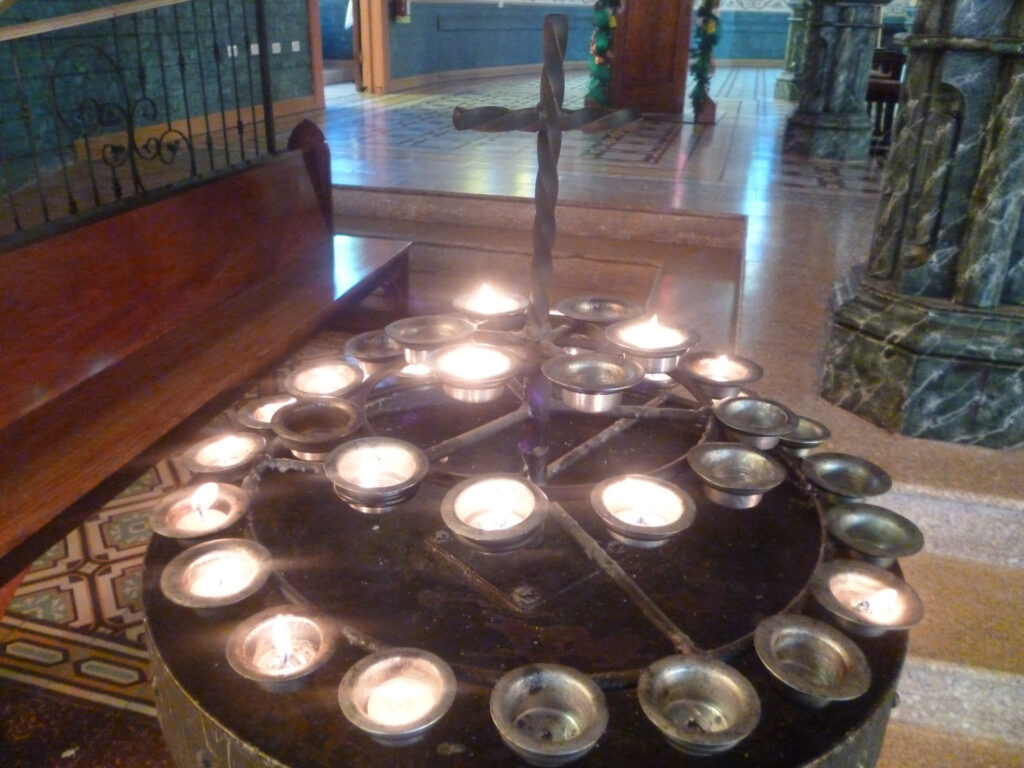 Fila di candele votive accese davanti a una parete di pietra, in una chiesa in penombra